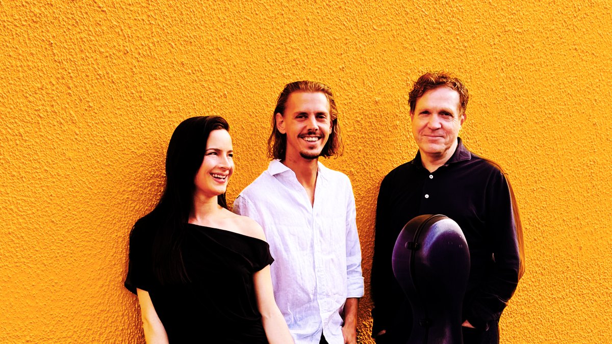 Three people, the musicians from Groove Cafe, stand closely together in front of a textured wall, painted a warm yellow. The three musicians wear black and white clothing to contrast the yellow wall behind them. The female presenting musician on the left leans against the wall with her right shoulder, and smiles warmly into the distance, laughing and having a good time. The two male presenting musicians on the right are smiling warmly at the camera, standing with their hands in their pockets. One musician has a cello case propped up in front of him.