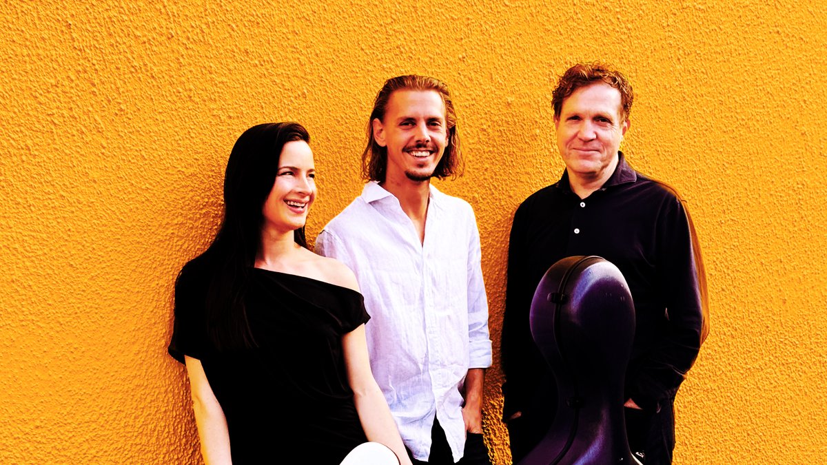 Three people, the musicians from Groove Cafe, stand closely together in front of a textured wall, painted a warm yellow. The three musicians wear black and white clothing to contrast the yellow wall behind them. The female presenting musician on the left leans against the wall with her right shoulder, and smiles warmly into the distance, laughing and having a good time. The two male presenting musicians on the right are smiling warmly at the camera, standing with their hands in their pockets. One musician has a cello case propped up in front of him.