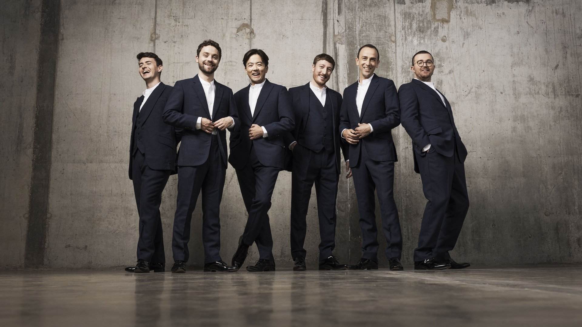 Six men known as The King's Singers stand in front of a plain background in matching navy suits. The King's Singers are all smiling and having a wonderful time, whilst standing next to one another in casual poses.