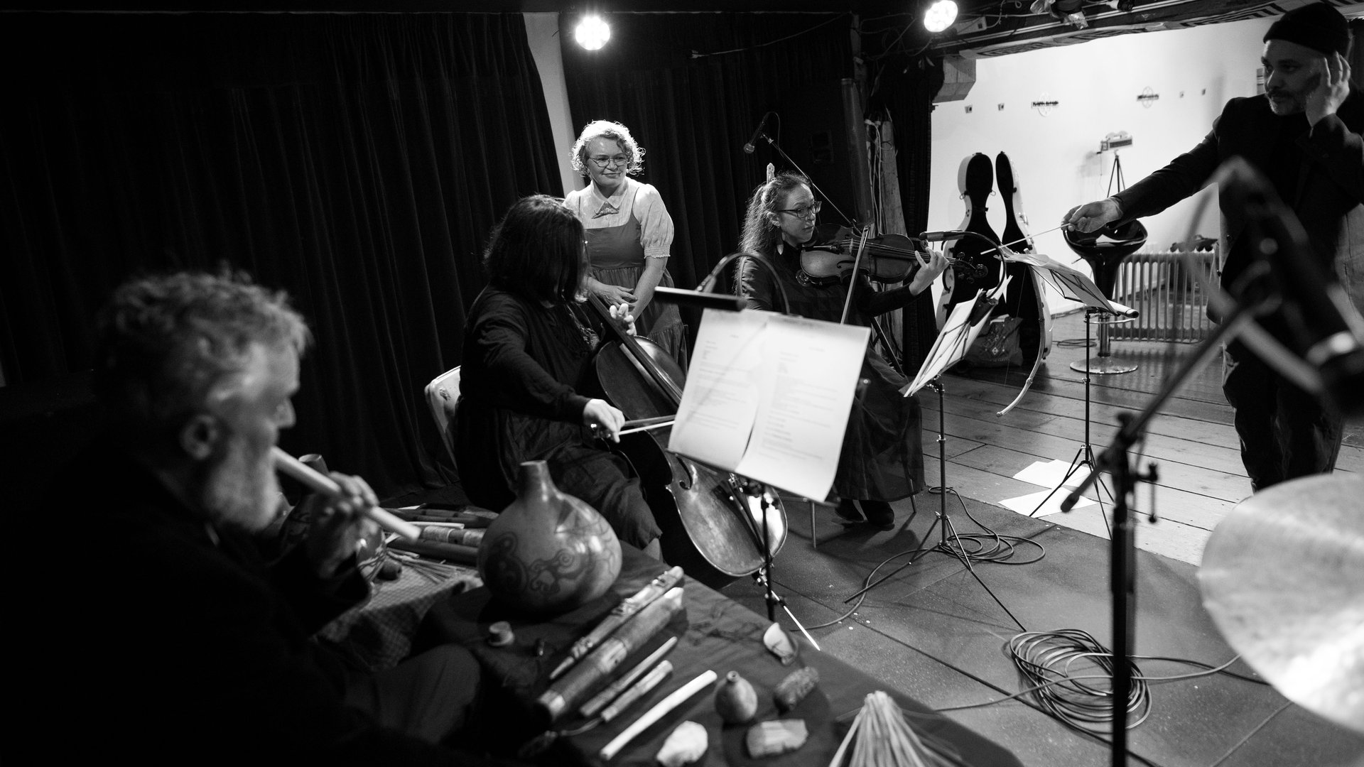 Three musicians are conducted by a leader standing before them. Of the three musicians, two are seated playing string instruments and one is seated behind a table displaying an array of indigenous instruments. A female-presenting person stands behind the musicians, admiring their work as she prepares to speak over the music.