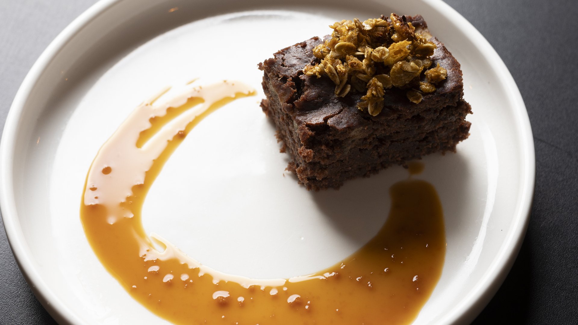A brown, crumbly square of cake, topped with various oats and nuts, is positioned on a white ceramic plate. To accompany the cake, a caramel-coloured sauce is artistically swirled around the plate to surround the cake.