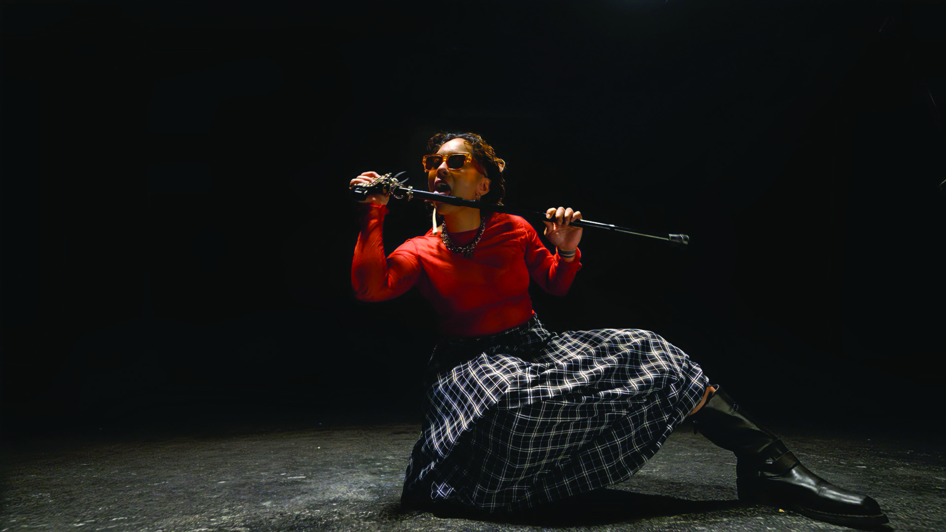 A female-presenting person in a striking red top crouches down in a huge, open black room. She wears orange sunglasses and holds a cane between her two hands as she licks it.