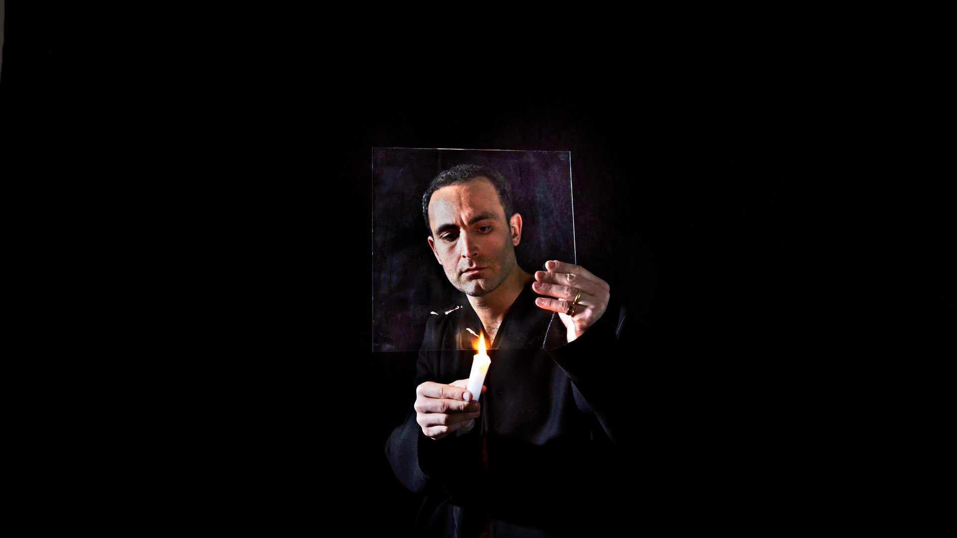 The solo actor for Nowhere stands in a pitch black room in pitch black clothes. The room lit only by a flaming, white candle in his hand. He holds a square glass pane in front of his face, holding the candle flame to the bottom. The man is reflective as he does this, staring closely at the flame.