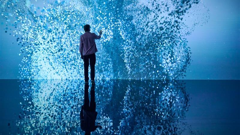 A person stands in an art gallery, fully surrounded by a projection of blue fragmented light. The person has a hand outstretched towards the art on the wall, and the fragments have parted slightly, as if they were interacting with the person. The light appears like water, movable and interactive.