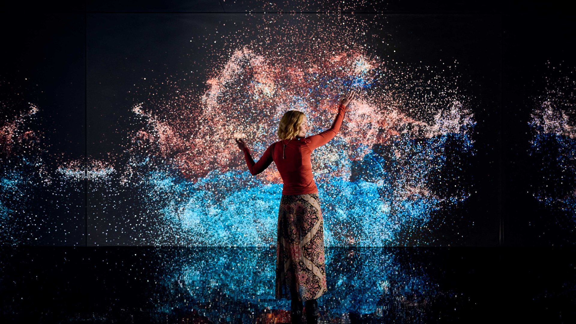 A pākeha woman in an orange long-sleeve and patterned skirt stands in front of a screen covered in an explosion of blue and orange stars on a black background. She faces the screen and gestures her hands up, amazed at the magical starry scene.