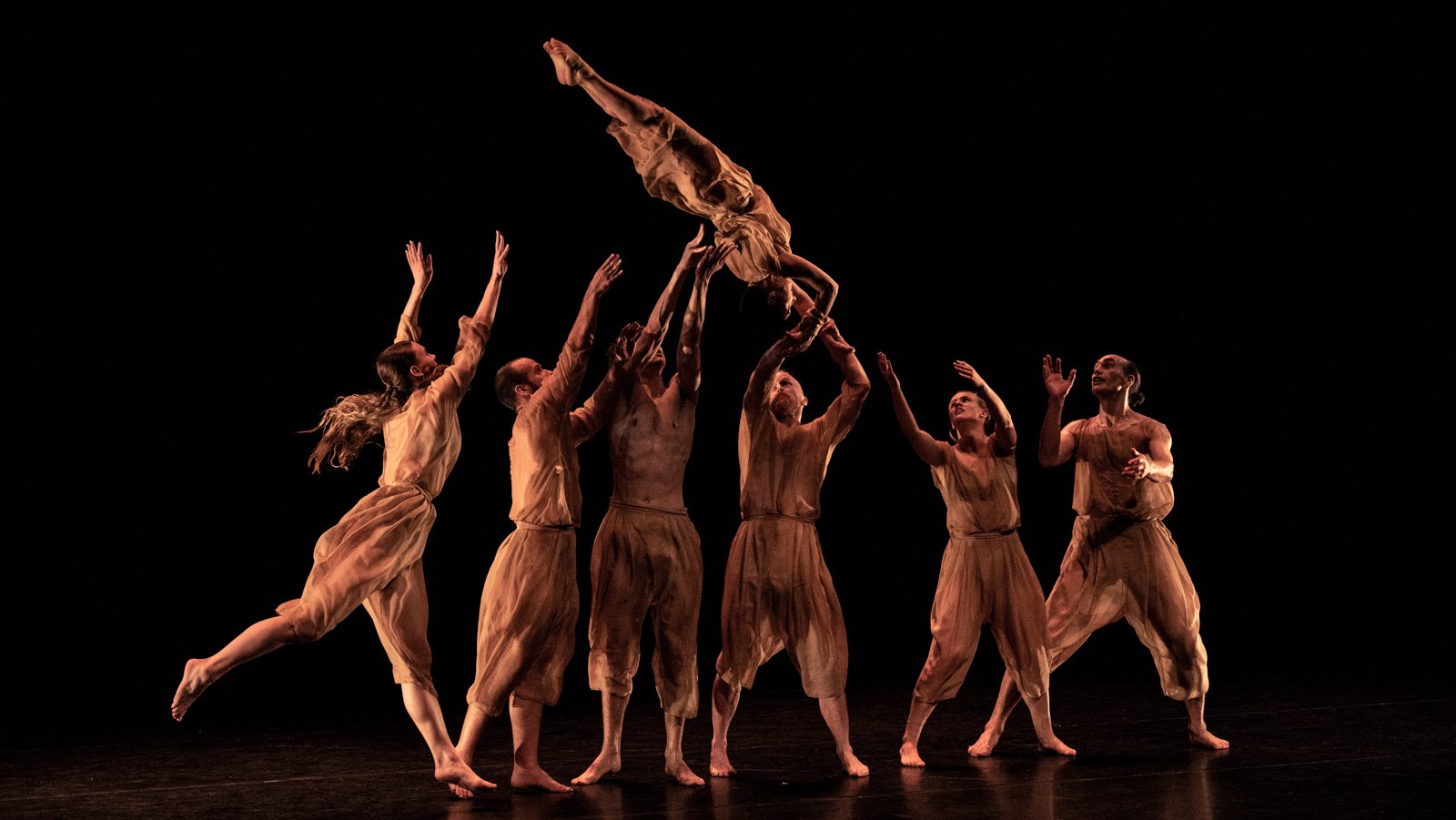Six dancers in 'Gloria' stand in a row, in loose, nude coloured clothing in front of a plain, black background. A seventh dancer in the same outfit is being tossed in the air - launched from the dancers on the right, to the dancers on the left. The dancer in the air holds themselves upside down in a strong plank position. All seven dancers show incredible strength and skill as they carry out this move.