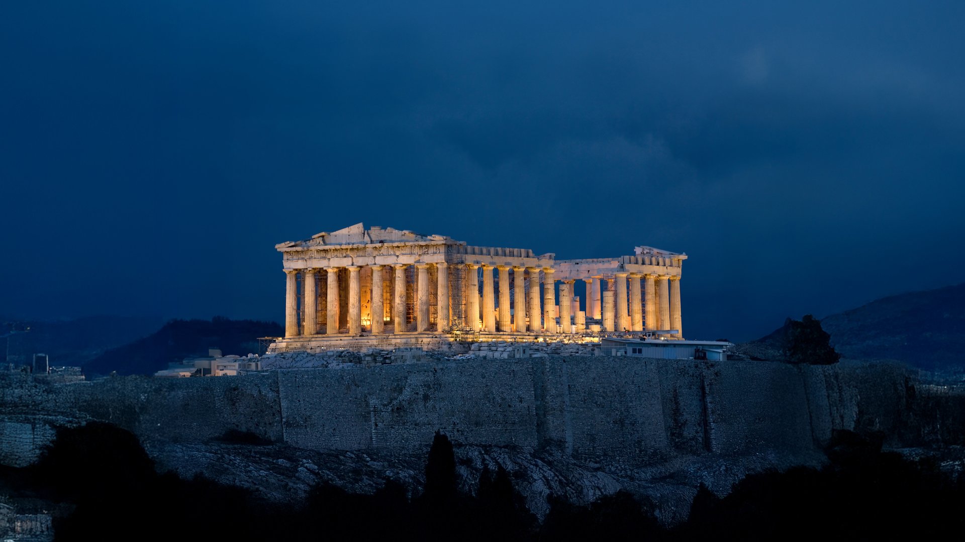 The Greek historical temple, the Parthenon, towers over the buildings surrounding the hill it stands on. The white marble building is lit with a warm yellow light, contrasting the blue, dusky, open air around it.