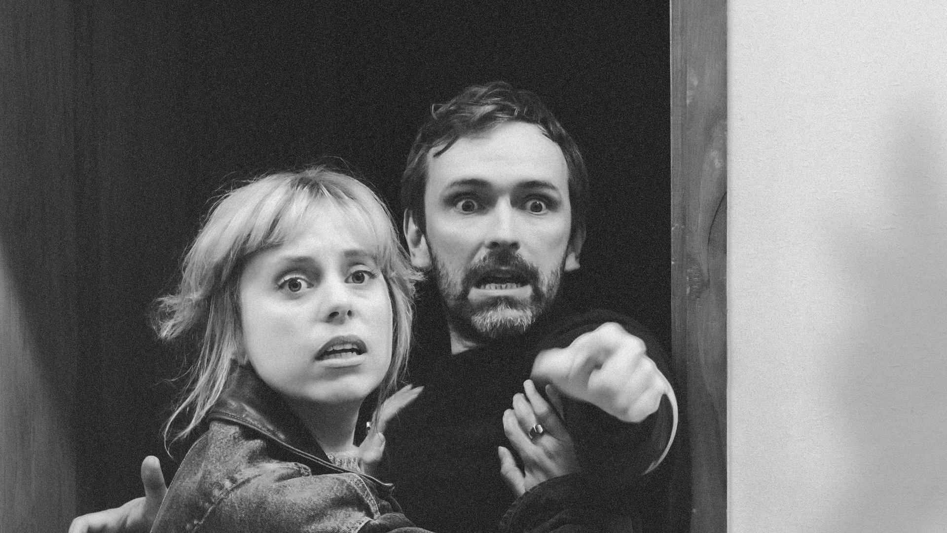 In this black and white image, two people look in fear at something off-camera. The one on the left is female presenting, has casually-tied up blonde hair, and wears a black leather jacket. The one on the right is a white male presenting person, with stubble, wearing a black top, and is pointing a finger towards something behind the camera. The female presenting person has her hands on the male’s chest. Both of their eyes have a lot of fear and urgency in them, as if whatever they’re looking at is coming for them.