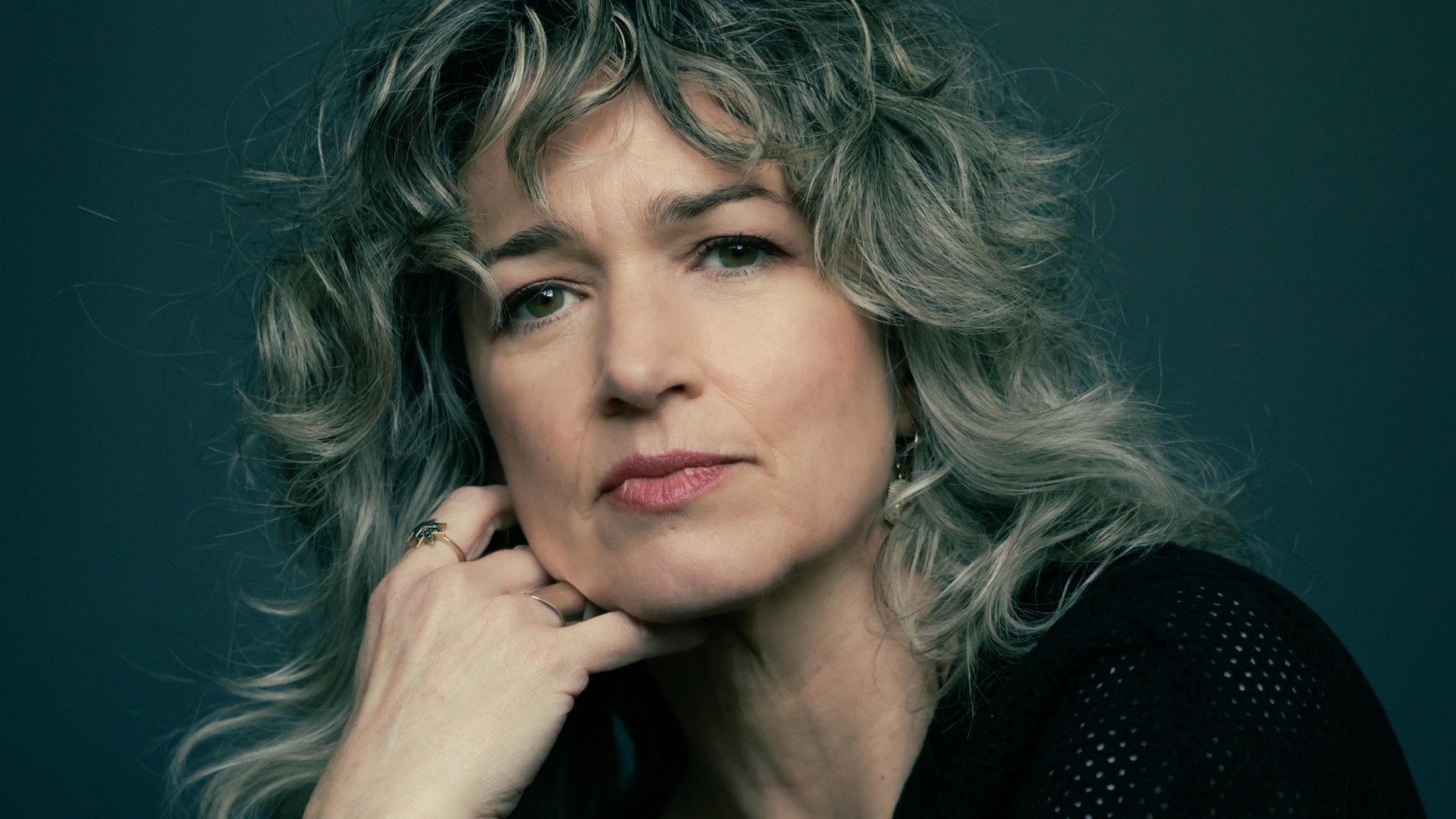 Julia Deans poses in a close up portrait. Her head rests on her hand as she stares at the camera with a powerful gaze. Her grey hair, clothes and the background behind her all create a calm atmosphere.
