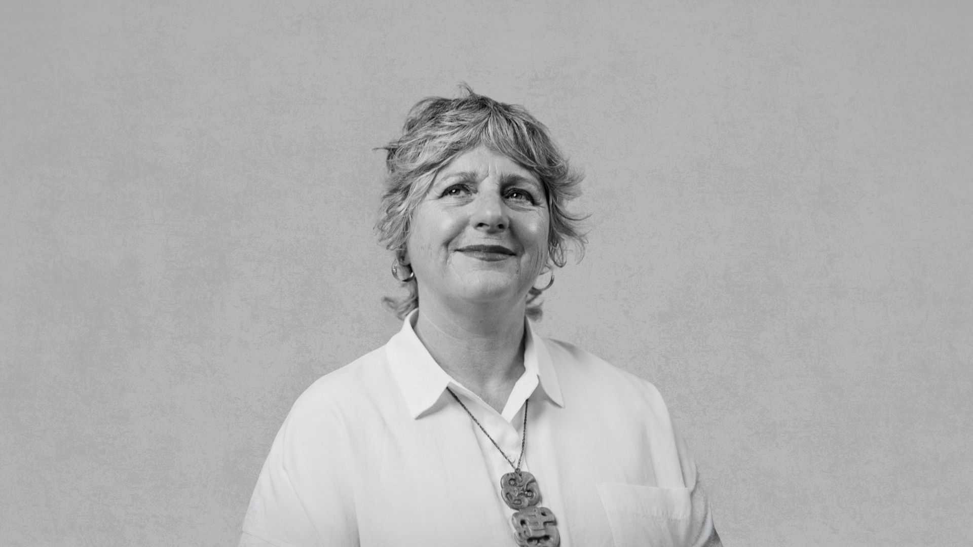 a black and white image of Dr Rachel Buchanan. She is looking toward the right and up at the sky. Her smile is soft and confident. Around her neck is a taonga, greenstone.