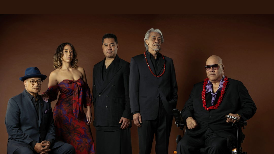Against a brown background, three performers stand in between two performers who are seated. The performer on the far right is Fonotī Pati Umaga, and he is in a wheelchair. Everyone in the image is looking directly at the camera with expressionless, but authoritative gazes. They are all dressed sharply, the men are in black suits, and the two men on the right have red lei around their necks. One of these men is Fonotī, and his lei Is larger, the other man is older with tidy, silver hair, and his lei is thinner. The woman is wearing a classy, off-shoulder, red-patterned dress. The colour of her dress matches the red of the lei. Her hand rests on the should have the seated man to her left. They look comfortable in each others company the way a family would.