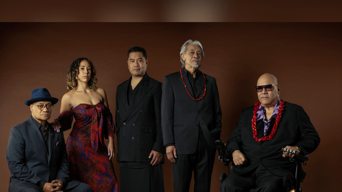 Against a brown background, three performers stand in between two performers who are seated. The performer on the far right is Fonotī Pati Umaga, and he is in a wheelchair. Everyone in the image is looking directly at the camera with expressionless, but authoritative gazes. They are all dressed sharply, the men are in black suits, and the two men on the right have red lei around their necks. One of these men is Fonotī, and his lei Is larger, the other man is older with tidy, silver hair, and his lei is thinner. The woman is wearing a classy, off-shoulder, red-patterned dress. The colour of her dress matches the red of the lei. Her hand rests on the should have the seated man to her left. They look comfortable in each others company the way a family would.