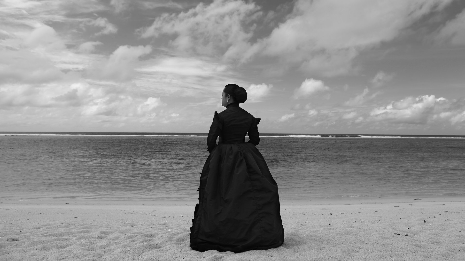 A female-presenting person in an all black, dramatic, long dress stares longingly out to the ocean