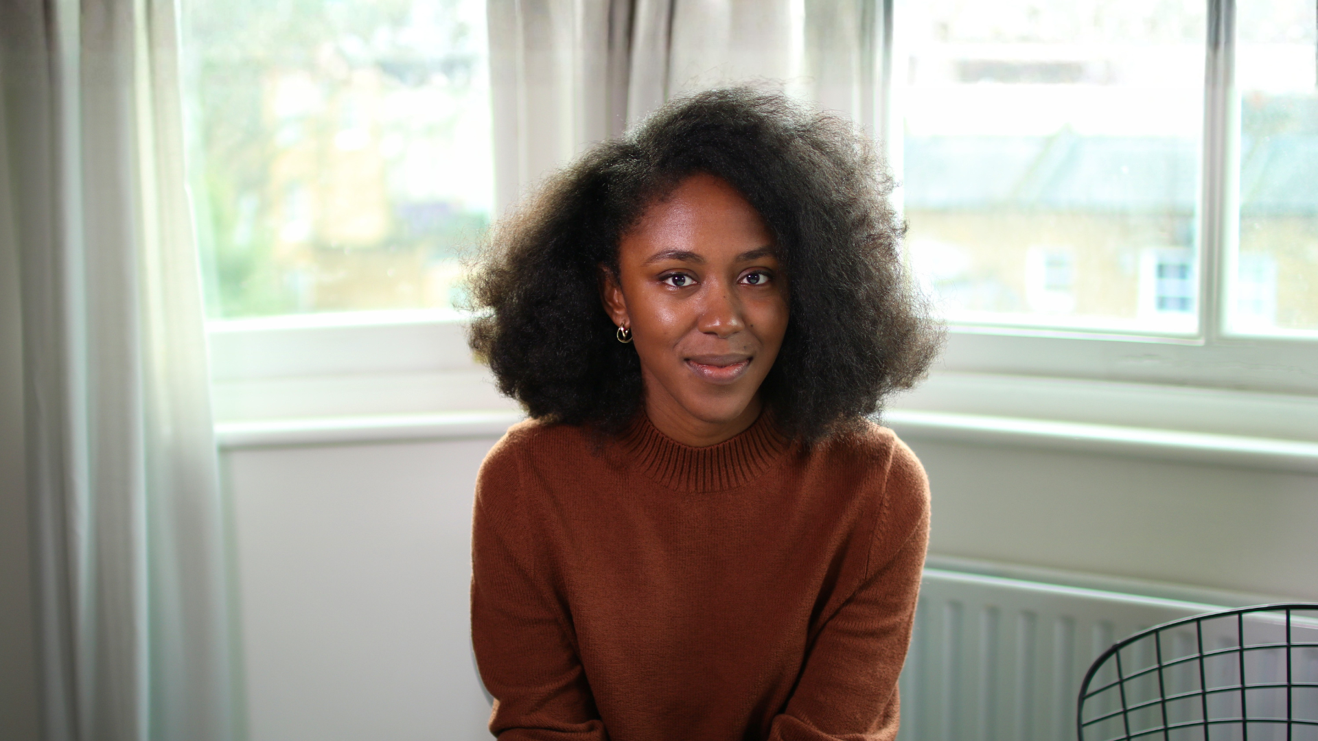 A female-presenting person with voluminous black hair and an earthy orange jumper smiles kindly. She is seated in a cosy white room.