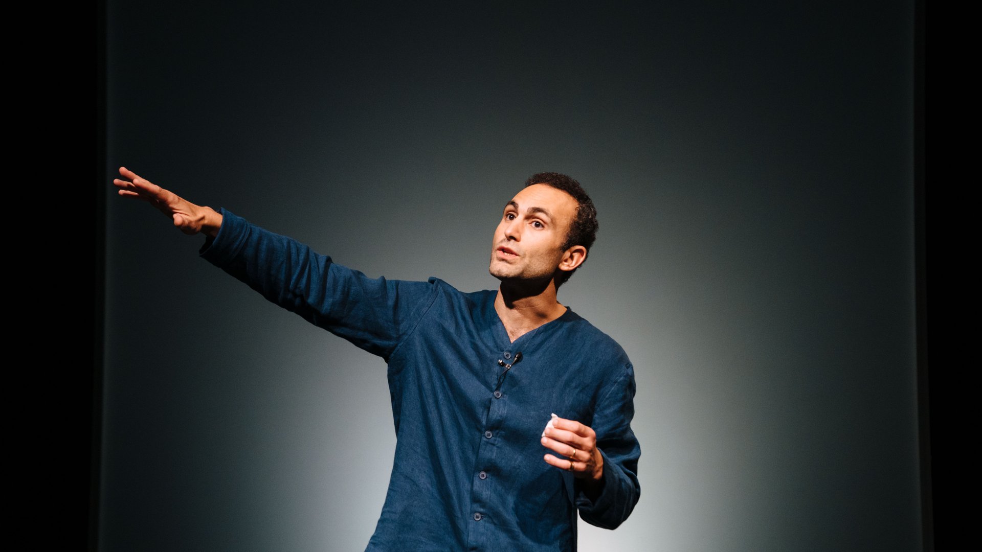 A male-presenting artist with short, black curly hair and a blue denim top with a microphone attached stands in front of a grey background. He gestures to his right with his hand as he speaks outwards.
