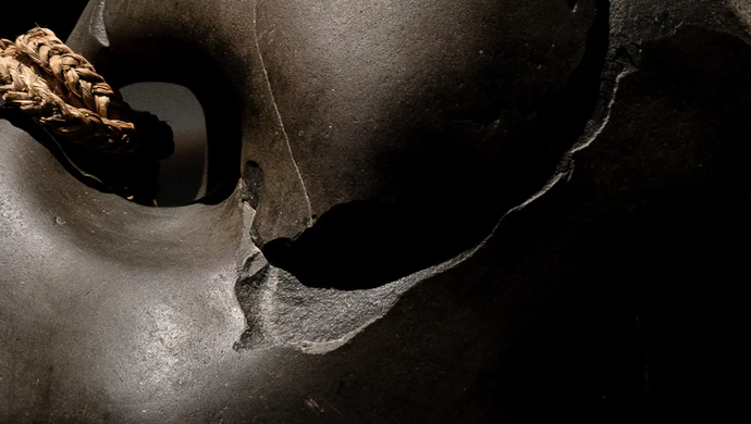 A smooth, dark brown sculpture rests around a bundle of woven rope. The sculpture has some significant chips and cracks in it.