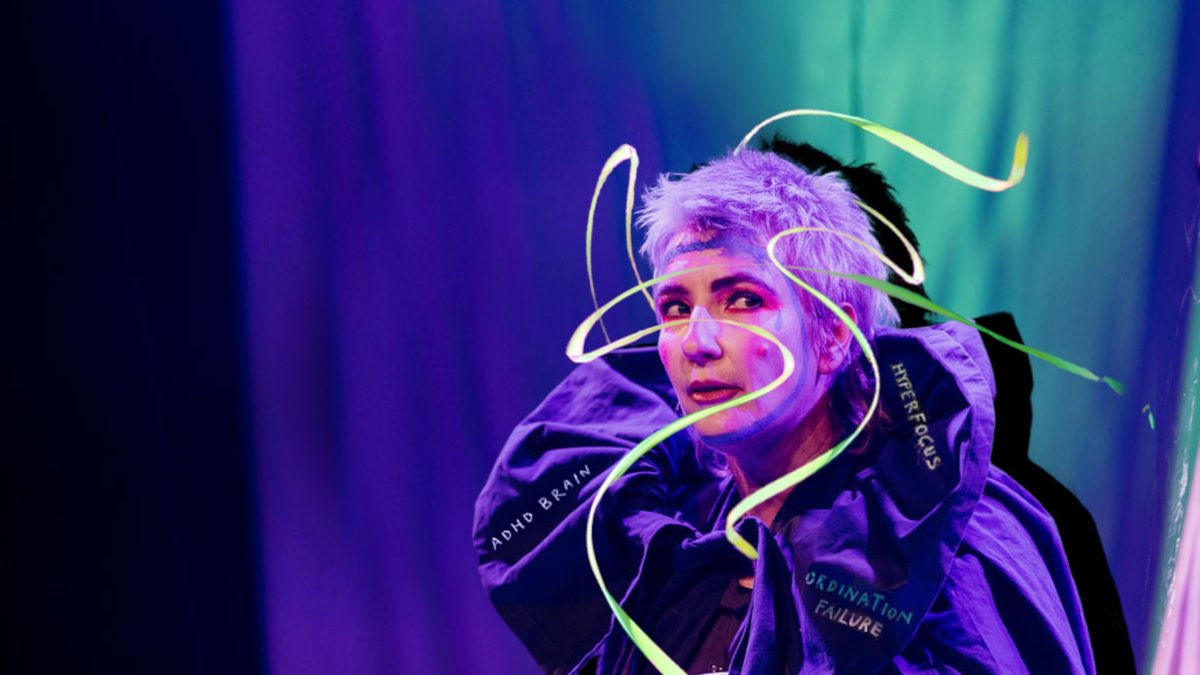 The performer for 'Speed Is Emotional' is hunched over and glancing cautiously to their left. Their clothes and the lighting reflecting onto their face is a royal, dark purple. The background is also purple, merging into a teal green towards the back right. The performer wears a dramatic, puffy purple cloak with collar. The collar has the phrases "ADHD Brain", "Hyper Focus" and "Ordination Failure" on it. The performer's hair is grey, short and spiky. They have green ribbons swirling around their face and over their head.