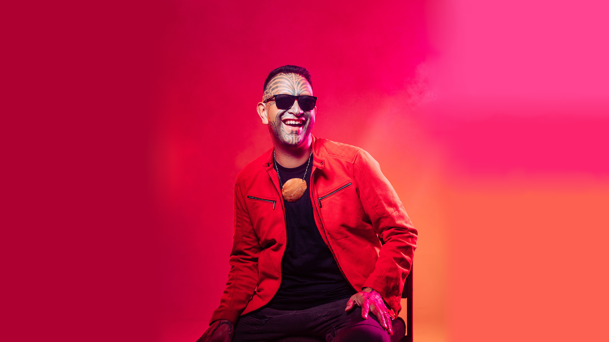 Rob Ruha sits on a chaior smiling and laughing, in a room with red, orange and pink lighting. He wears a red jacket to match his surroundings and black sunglasses covering his eyes. A wooden, carved, māori taonga sits on his chest, hung by a necklace chain. He has moko (māori tattoos) on most of his face.