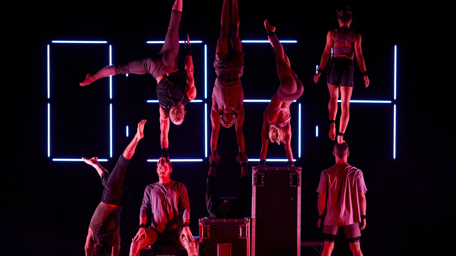 Eight performers are bathed in pink light in front of a digital clock screen, lit up with the numbers 0354. The performers appear to be acrobats, as there are four standing, and three doing handstands on top of the standing performers, and one standing on one of the performers heads. They wear grey, athletic, casual clothing. They all appear very athletic and agile. It is as if the image implies variation, and excitement, as the performers are all doing something slightly different, but all very impressive.