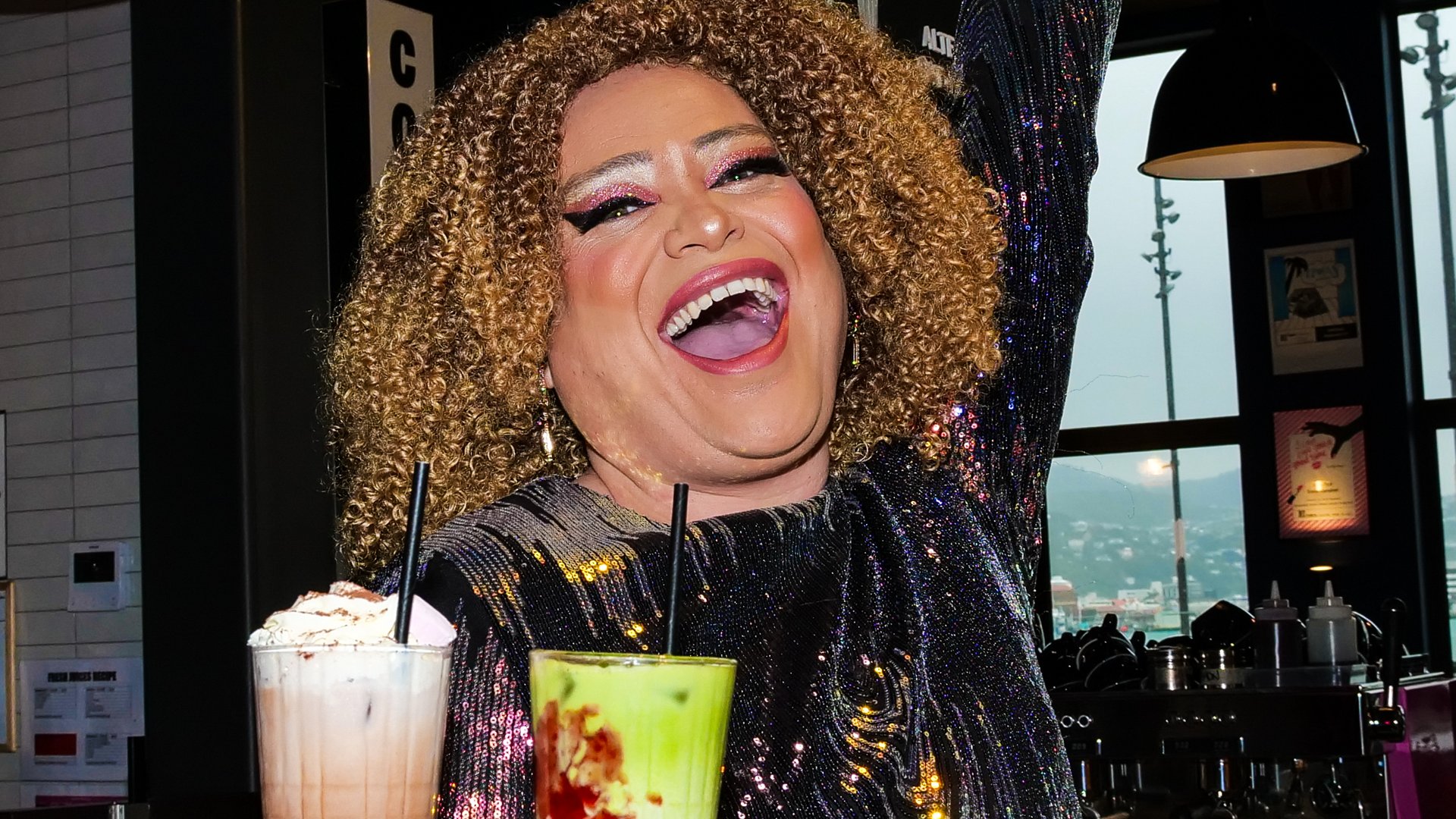 A Pasifika drag queen smiles widely and exuberantly while holding a tray balancing two interesting looking drinks. You can see that she is in a restaurant or café, as there is a coffee maker on a bench behind her. Her hair is neat and very curly, and her makeup includes thick eyeliner, sparkly pink eyeshadow, highlighted brow bones, blush, and cherry red lips. She is wearing a dark, sparkly top which catches light from the near, overhanging lamp. One of the drinks she holds is green with chocolate brown swirls and a straw; The other drink is white with caramel-coloured swirls, whipped cream on top, and a straw.