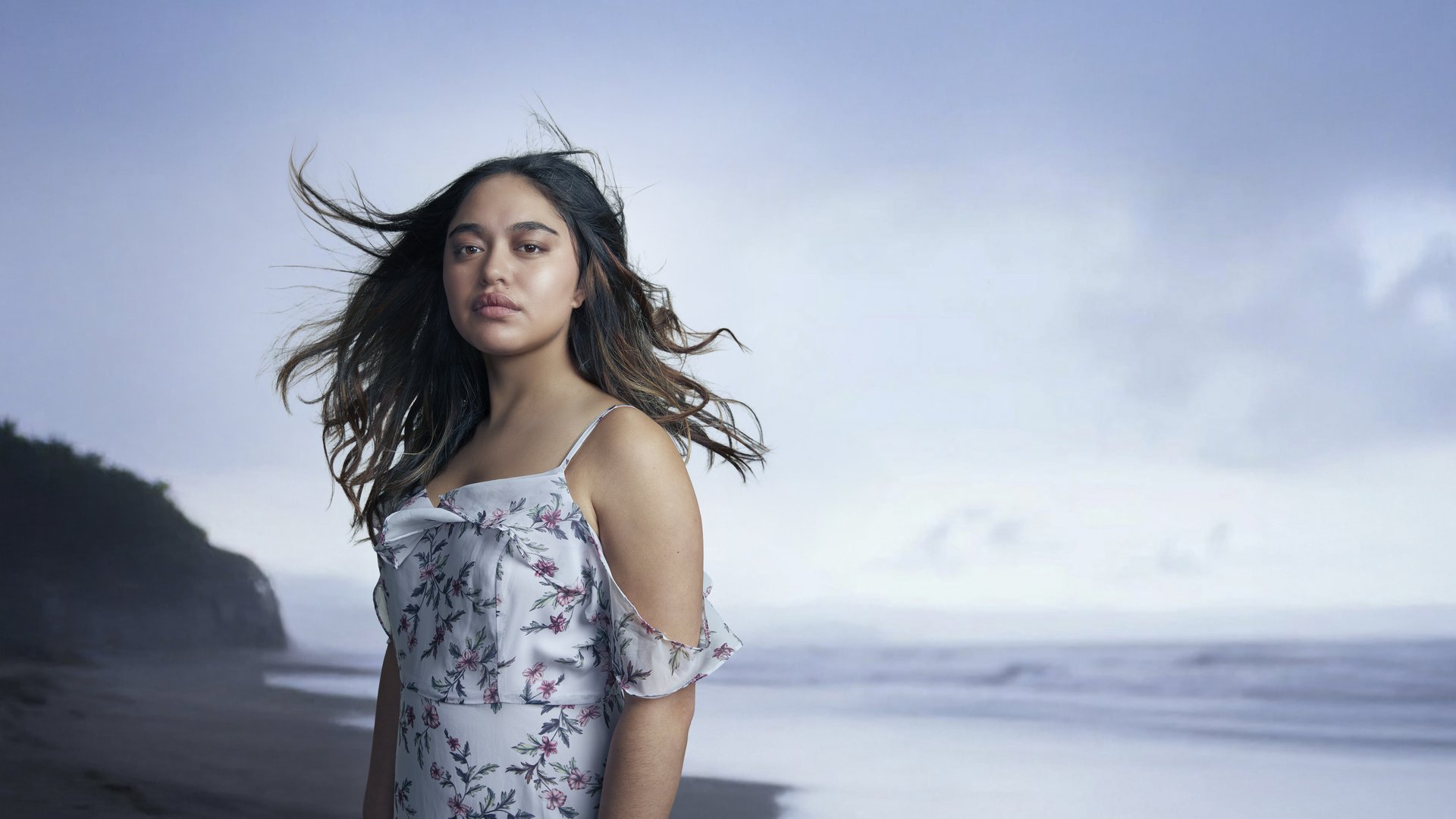 A young woman with tanned skin and long, brown hair stands firmly on an isolated beach. The beach in the far background features black sand with dusky skies and crashing waves. The woman's hair is blowing with the wind, yet she still stands unflustered.