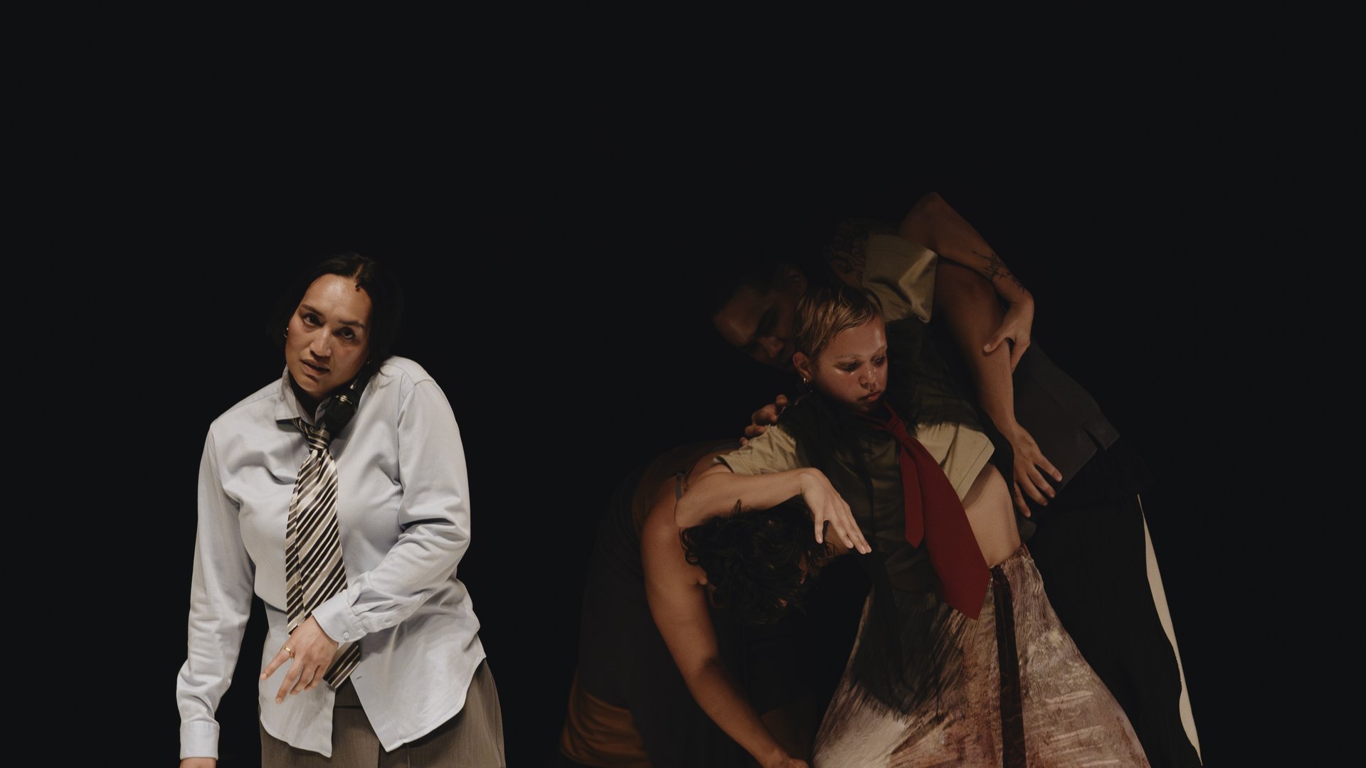 Three dancers are brightly light amidst a background of darkness. One stands apart from the other two on the left of the image. This female-presenting person wears a long sleeve, light blue shirt with a black and white tie. Her eyes are fixed on the camera as she is in the middle of a dance move. Her body is fully facing the camera, but her left shoulder is raised to her ear, and her head is slightly tilted towards it. On the right, and slightly behind the first performer, two figures are intertwined in a dance move. Their costumes are earthier in tone, and the one in front has a visible red tie. They are not looking at the camera, seemingly entranced by the dance move. The one behind them has their arms reached over their shoulders. The one in front has their elbows raised, almost as if they are pushing them away.