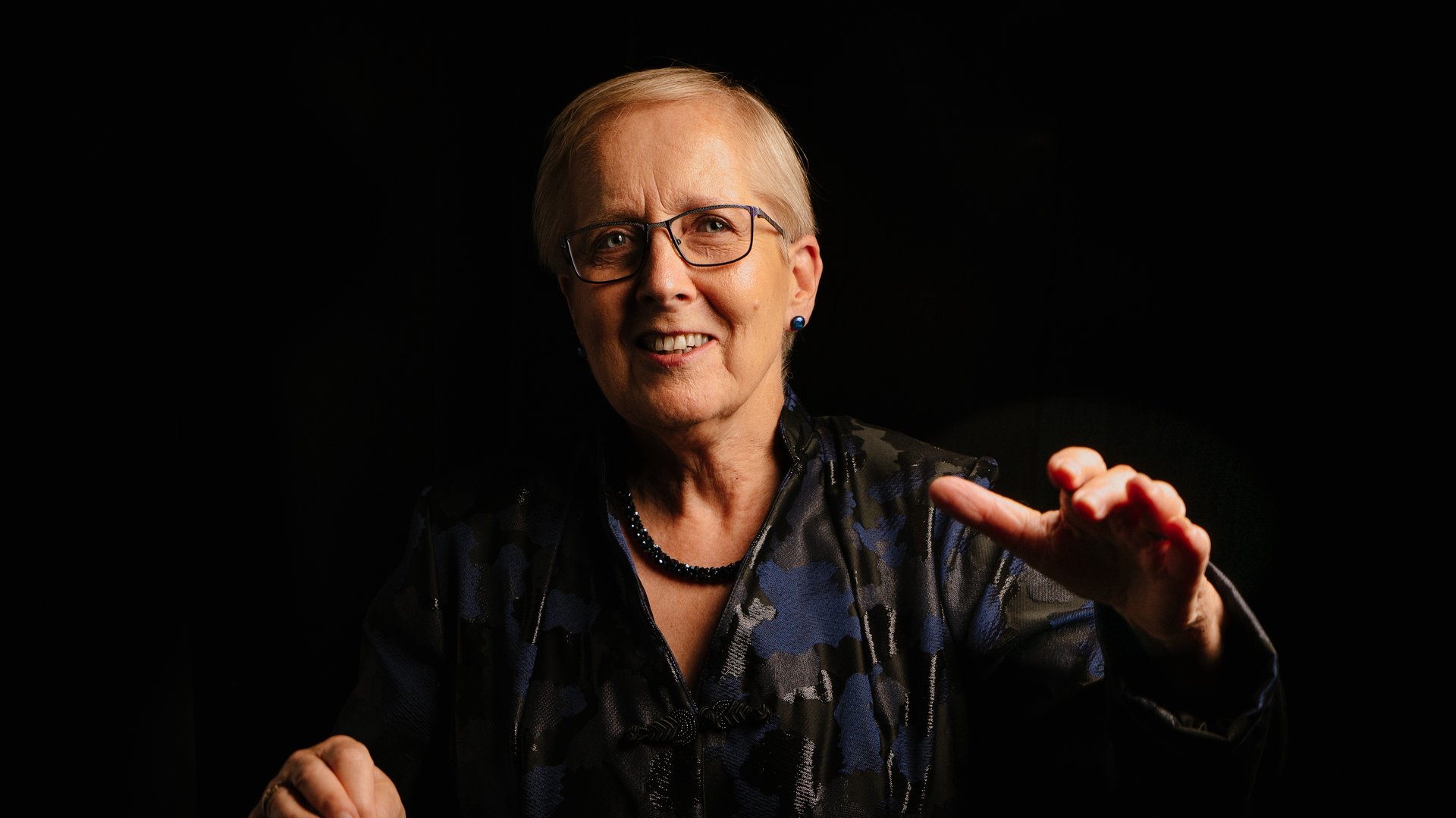 This portrait of Karen Grylls strikingly distinguishes her from a dark background. She is warmly lit and tidily dressed. Her hand is raised toward the camera, as if she is gesturing while speaking, or perhaps conducting. The expression on her face evokes a quiet joy, as she has a small smile and bright eyes. She wears a black and blue top, a thick black necklace, blue stud earrings, and black glasses with thin lenses.