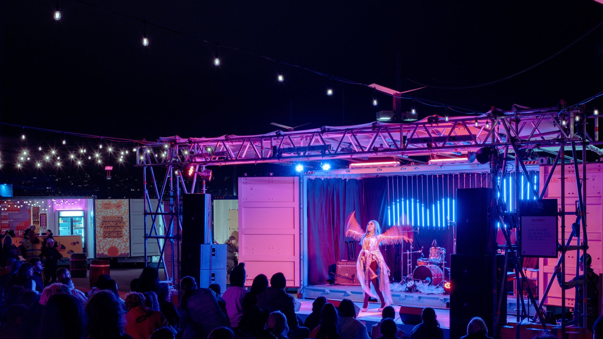 An outdoor stage at night time is lit with pink and blue light, and a few rows of fairy lights streaming from the from stage awning, over the audience. There is a lone, female presenting performer caught in the midst of waving her arms, and the image captures the movement with motion blur. The audience watching is full and encapsulated in the performance. To the left of the stage, there is a food truck, lit up. The stage appears to be a converted shipping container. The image as a whole conveys spontaneity and wonder.