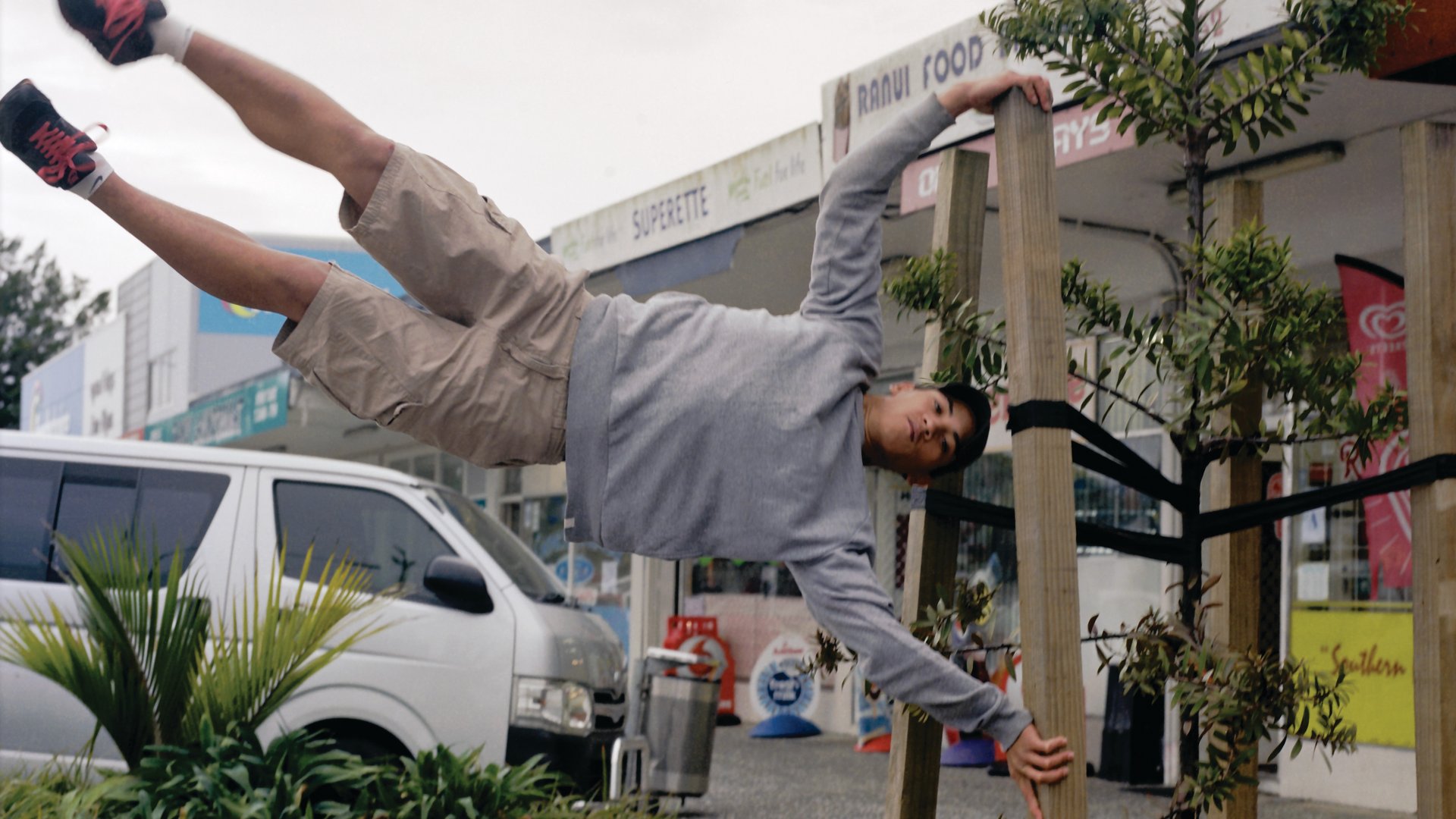 A young, male-presenting person holds himself sideways and in the air, hanging off a wooden post. He concentrates deeply as he uses great strength to hold himself off the ground.
